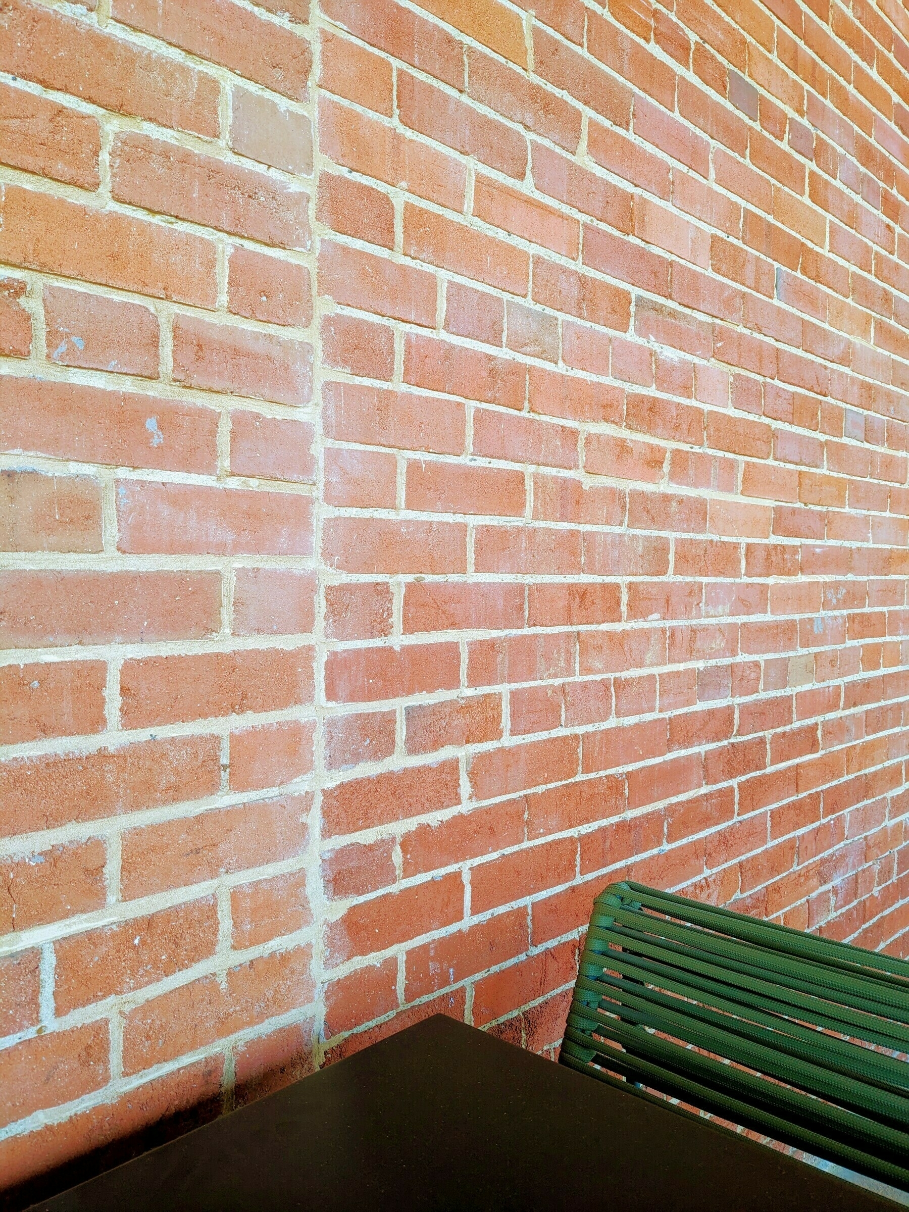 Pinkish-red brick wall with a vertical grout seam in white-cream beside the corner of a black table and green chair. 