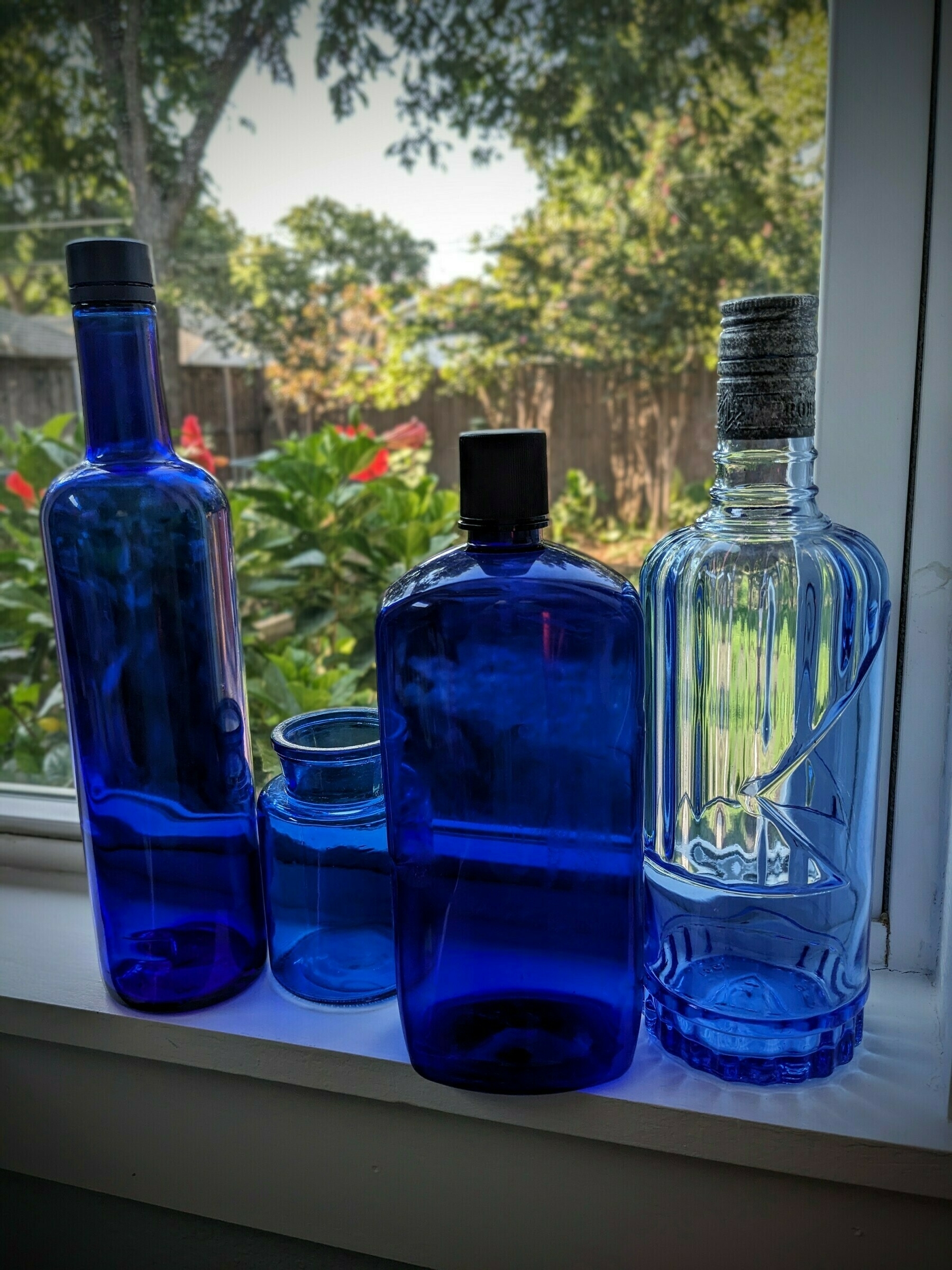 Four deep blue bottles of different intensities, sizes, and shapes on a white windowsill with the outdoors in the background. 