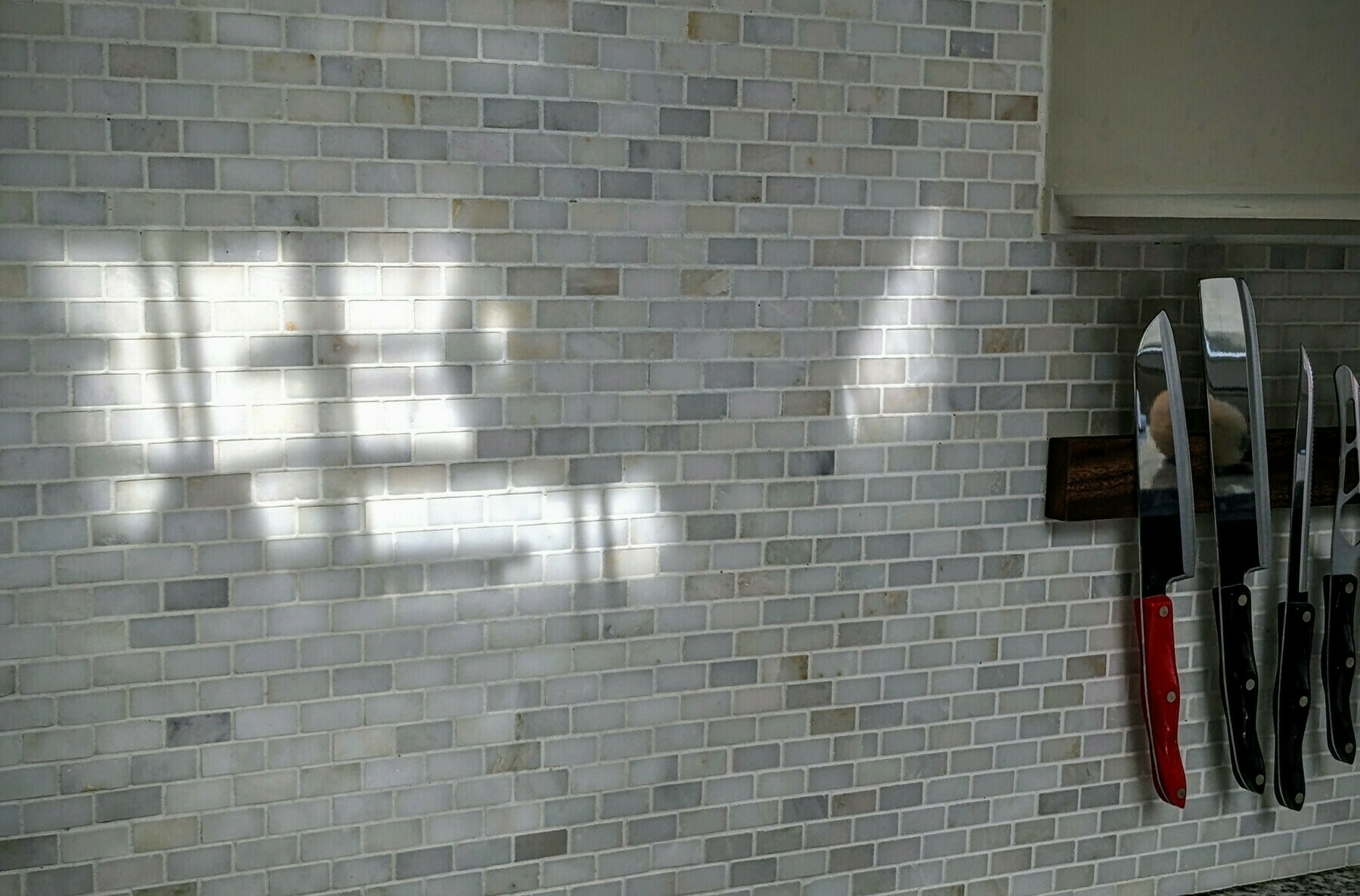 Tiled kitchen backsplash with interesting indeterminate shadows and a magnetic knife rack on the right, with cleavers and knives, including a red-handled cleaver, which is the only strong color in a mostly black, white, and gray image