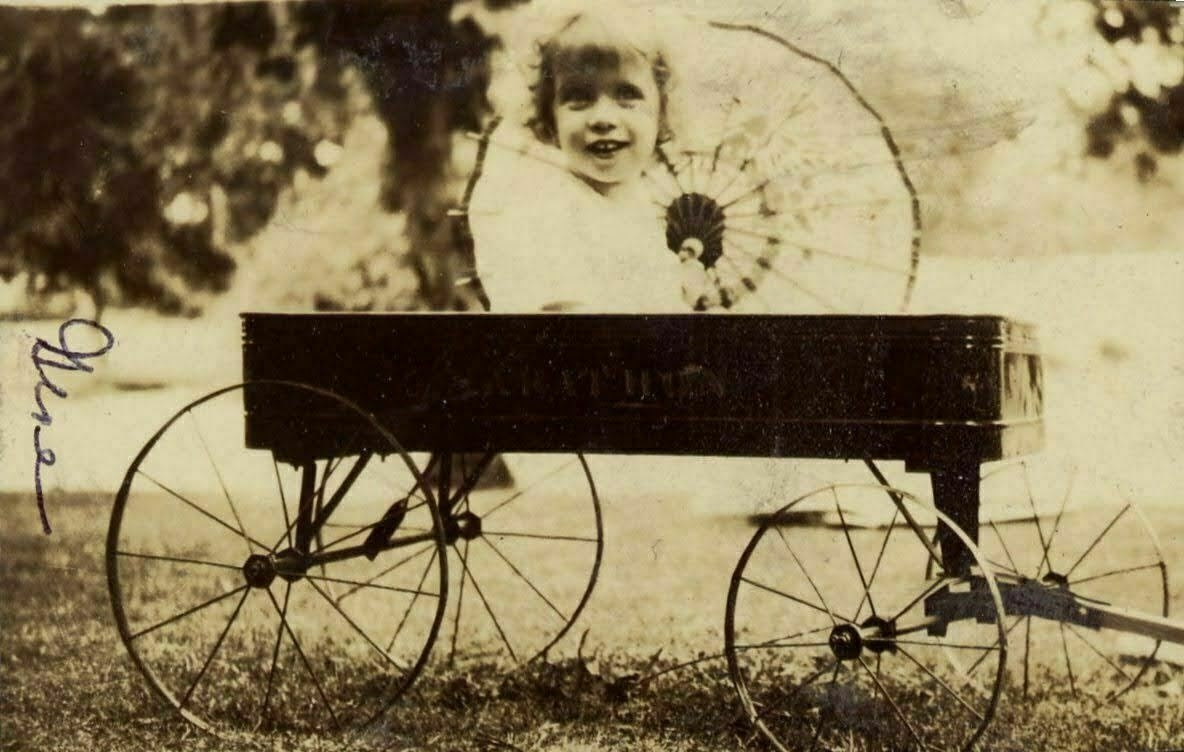 Old sepia-toned photo of a happy little girl in a wagon holding a parasol.
