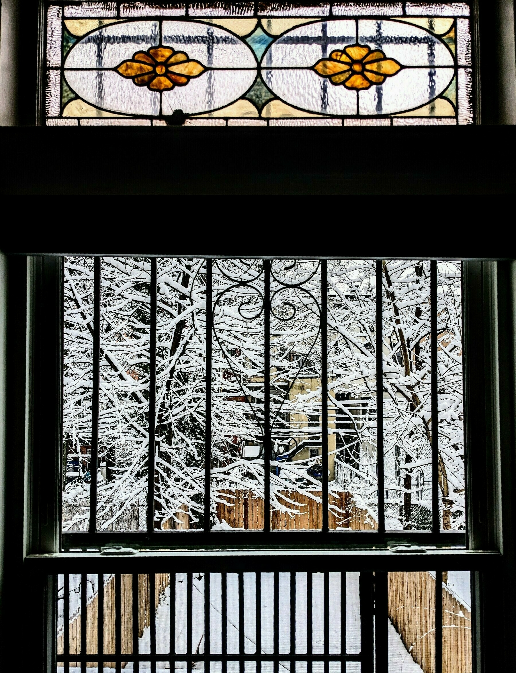 Snow-covered tree branches seen through a window with a grate and stained-glass panels